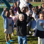 Group of smiling children outside on a sunny day with their hands raised toward the camera.