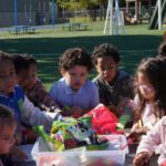 Children of diverse backgrounds gathered around a table outside, sorting colorful snacks from a plastic bin, with a smiling adult nearby.