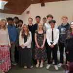 Group of diverse teens posing together in a decorated hall with holiday wreaths and a Christmas tree, some in dresses and ties, smiling for a photo.