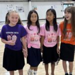 Four girls stand in a gym holding hands, wearing matching colorful shirts that read 'Twinning With' and feature cupcake graphics.