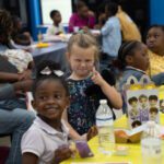 Smiling young girls at a birthday party table with snacks and drinks, thumbs up gesture in the foreground.