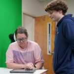 Woman in a pink shirt and glasses sits at a table, focused on a tablet, as a young man in a blue hoodie stands beside her in a room with a green screen backdrop.