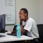 Young man in a white t-shirt sits at a computer desk in a classroom, with a blue water bottle on the table.