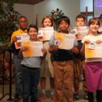 Eight smiling children stand on a church stage, each holding an award certificate for achievement. A cross and floral decor are in the background.