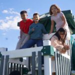 Four kids on a green-and-white playground structure, enjoying a sunny day on a slide platform.