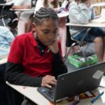 Student in a red shirt working on a laptop at a classroom desk while peers study nearby.