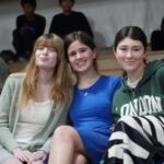 Three young women sitting on gym bleachers, smiling at the camera.