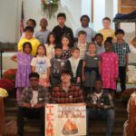 Group of diverse children and teens posing on church stair steps for a team photo; in front are three young adults holding a sign that reads 'TEAM SMORES' with a campfire illustration.
