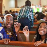 Two smiling girls sitting in a church pew, each holding a small football in front of a crowded hall.