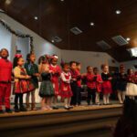 Group of children singing on a church stage during a Christmas program, dressed in festive outfits with holiday decor and a cross in the background.